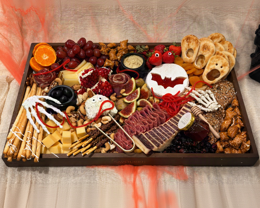 A Halloween-themed charcuterie board arranged on a dark rectangular tray, featuring cheeses, cured meats, crackers, grapes, an orange slice, pretzels, nuts, and spooky decorations like skeleton hands, a bat-shaped cookie, and red spiderwebs on a beige fabric background.
