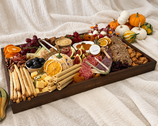 A wooden charcuterie board filled with cheeses, cured meats, crackers, grapes, citrus slices, nuts, and small gourds, styled on a cream fabric backdrop with pumpkins around it.