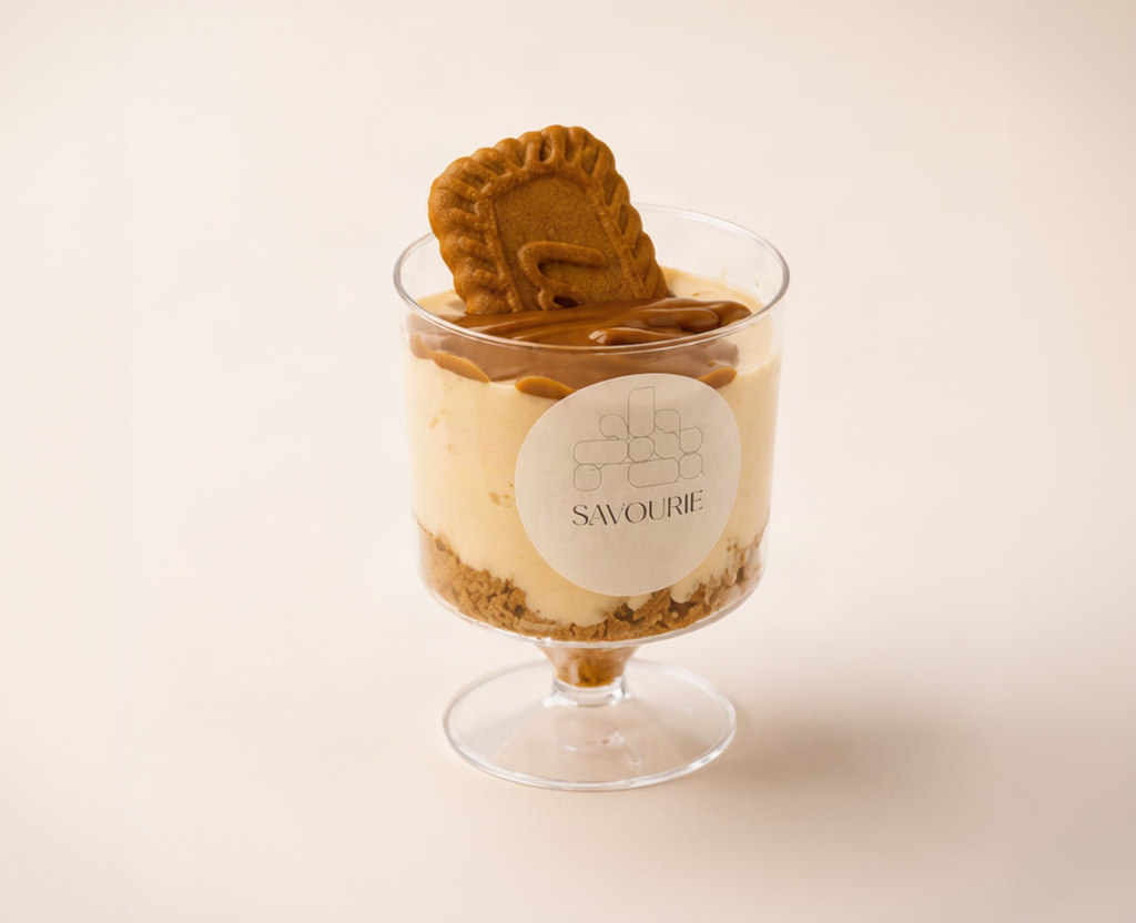 A dessert parfait in a clear glass pedestal cup with a round white label reading 'SAWOURI', layered with cream, chocolate sauce, biscuit, and a cookie on top, set against a warm cream background.