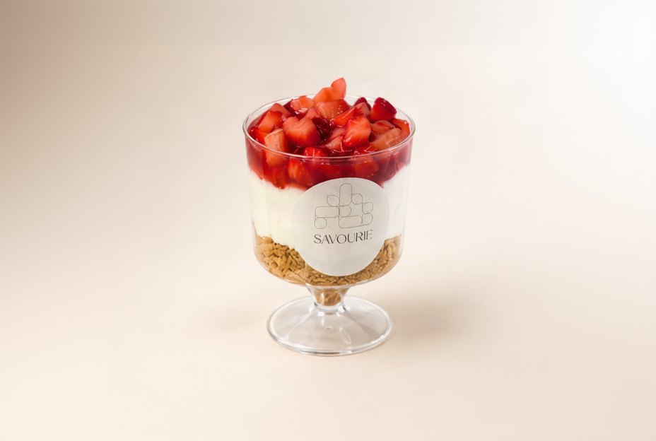 A glass parfait dessert with a biscuit base, white cream, sliced red strawberries on top, and a white round label reading SAVOIRIE in the center, set against a warm beige background.