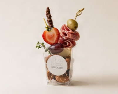 A clear plastic cup filled with assorted charcuterie snacks, garnished with a strawberry slice, a chocolate-covered stick, a green olive, sliced salami, crackers, and a round white Savourie logo label on the front, set against a plain beige background.