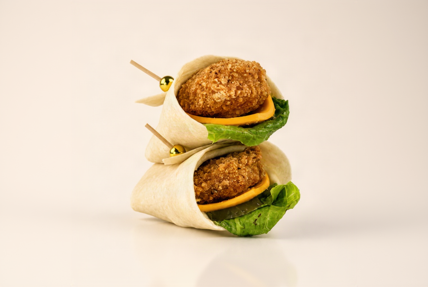 Two mini wraps stacked on a light beige background, each filled with a golden-brown fried patty, a slice of yellow cheese, and green lettuce, secured with wooden skewers.