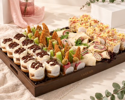 A large wooden serving tray filled with assorted finger foods and appetizers, including mini sandwiches, wraps, fried items, and drizzled chocolate sauce, styled on a cream fabric surface with soft shadows and decorative flowers in the background.