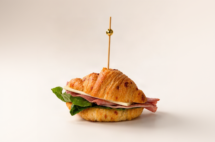 A croissant sandwich with ham, cheese, and green lettuce, secured with a wooden toothpick featuring a gold ball ornament, set against a plain light beige background.