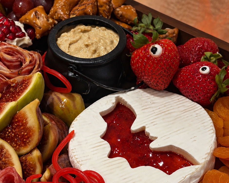 A Halloween-themed charcuterie board filled with spooky treats, including a round cheese wheel with a bat-shaped fruit filling, red licorice, strawberries with googly eyes, dried apricots, pretzel rods shaped like bones, and a small bowl of dip.