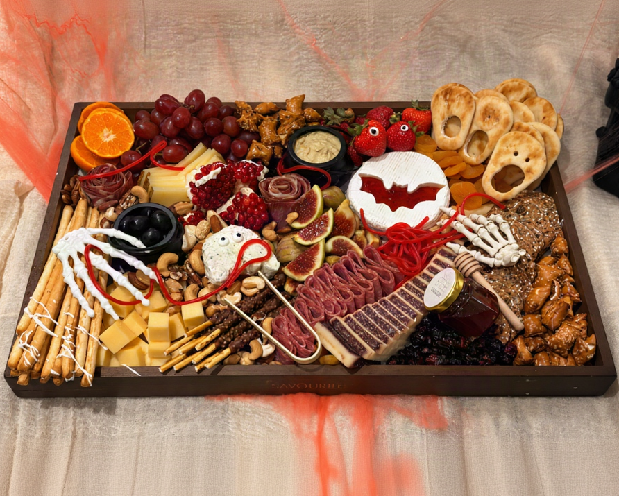 A Halloween-themed charcuterie board arranged on a dark rectangular tray, featuring cheeses, cured meats, crackers, grapes, an orange slice, pretzels, nuts, and spooky decorations like skeleton hands, a bat-shaped cookie, and red spiderwebs on a beige fabric background.