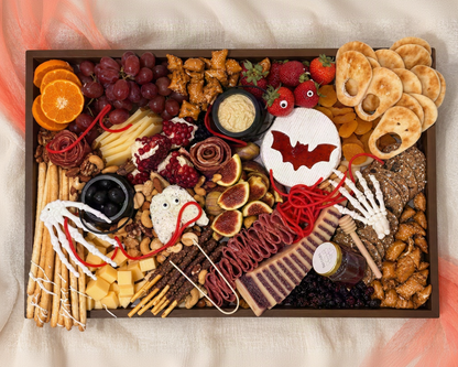 A Halloween-themed charcuterie board arranged in a rectangular wooden tray, featuring spooky snacks like pretzel sticks, cheese, crackers, grapes, figs, candy corn, gummy bears, and a bat-shaped treat, styled on a beige fabric background with orange and black fabric accents.