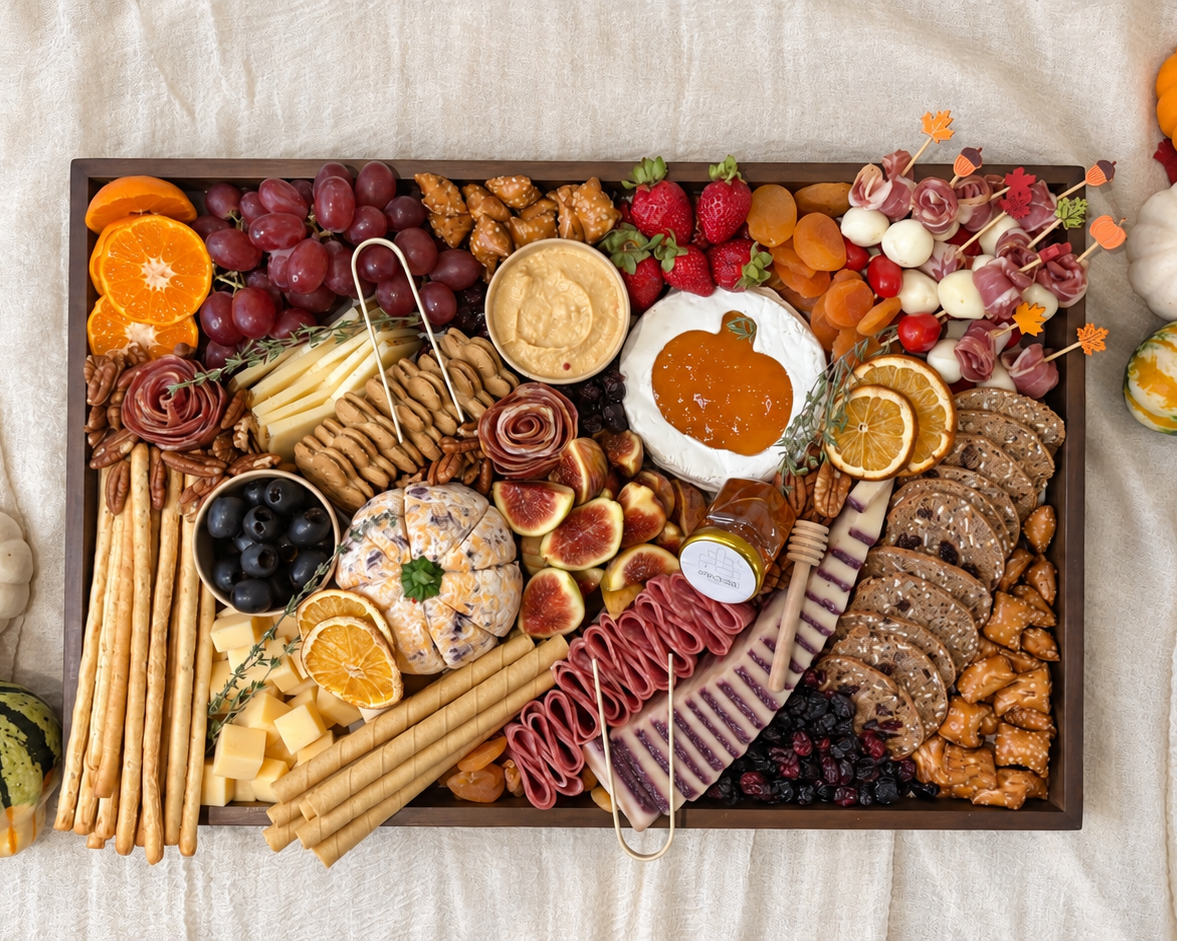 A large charcuterie board arranged in a dark rectangular tray, filled with cheeses, crackers, fruit, cured meats, nuts, and dips, styled on a cream fabric background with small pumpkins around it.