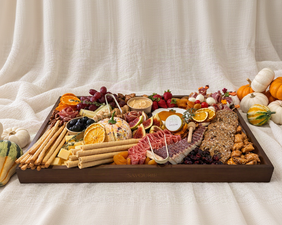 A large wooden charcuterie board filled with cheeses, crackers, fruit, nuts, and cured meats, styled on a cream fabric backdrop with small pumpkins around it. The board is branded with the name SAVOURIE on the front edge.