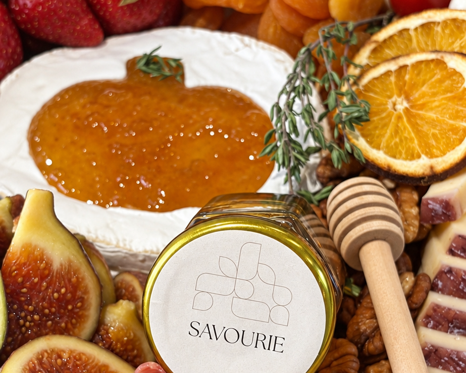 A close-up styled food spread featuring a jar labeled SAVOURIE with honey on the lid, surrounded by sliced salami, prosciutto, brie cheese, figs, dried apricots, strawberries, cherry tomatoes, orange slices, and a wooden honey dipper on a wooden board.