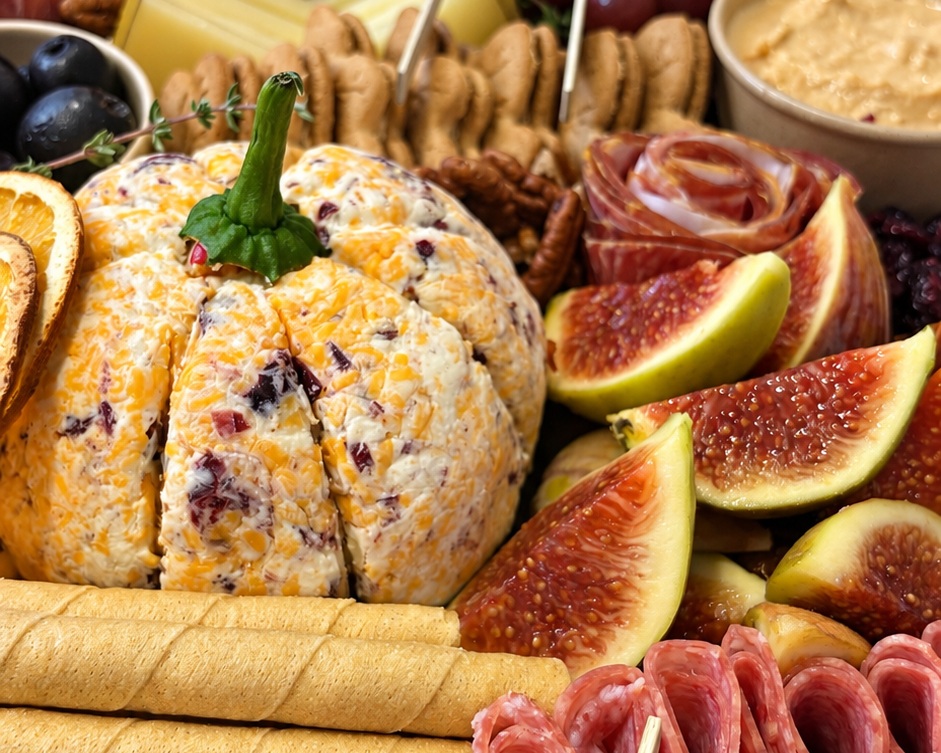 A close-up of a charcuterie and cheese board arranged on a white fabric surface, featuring sliced cheeses, rolled salami, cured meats, grapes, figs, dried fruit, crackers, and dips.