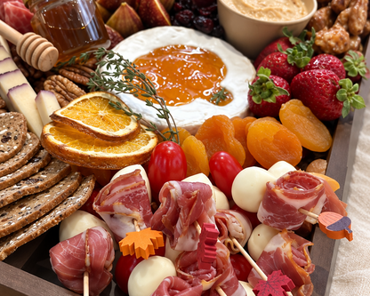 A large wooden charcuterie board filled with sliced meats, cheeses, crackers, fruit, dried apricots, nuts, and small skewers on a beige fabric surface.