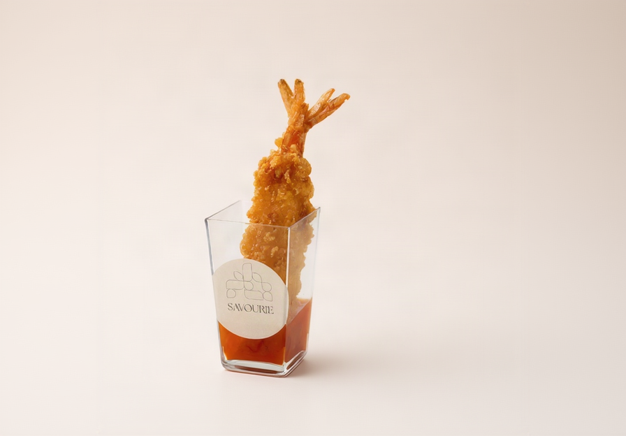 A golden-brown tempura shrimp skewered vertically in a clear square glass container filled with red sauce, set against a solid light beige background. The glass features a white circular label with the brand name 'SAVOURIE'.