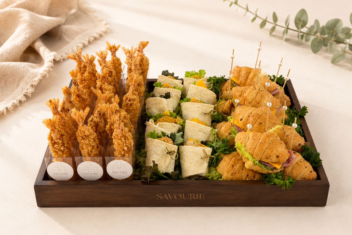 A wooden serving tray labeled 'SAVOURIE' filled with assorted appetizers, including golden tempura, wrapped sandwich rolls, and croissant sliders with toothpicks, set on a light beige surface with a folded cloth and eucalyptus sprig in the background.