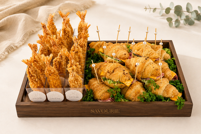 A wooden serving tray labeled SAVOURIE, filled with assorted appetizers including golden shrimp tempura, croissant sandwiches on a bed of greens, and three small dipping sauces, styled on a light surface with a beige cloth and eucalyptus branch.