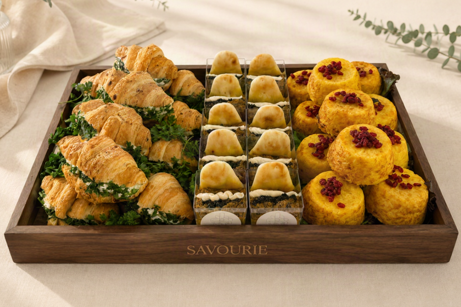 A wooden serving tray labeled 'SAVOURIE' filled with assorted pastries and breads, arranged on a cream fabric background with soft greenery accents. The tray contains croissants, small filled pastries, rectangular sandwich pastries, and round golden-yellow baked goods topped with red berries.