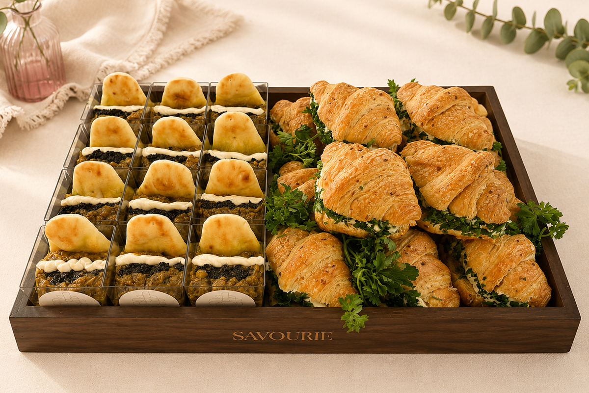 A wooden tray of assorted savory pastries and mini tarts, arranged on a cream fabric background with decorative greenery and a small pink glass vase. The tray is branded with the word 'SAVOURIE' on the front.