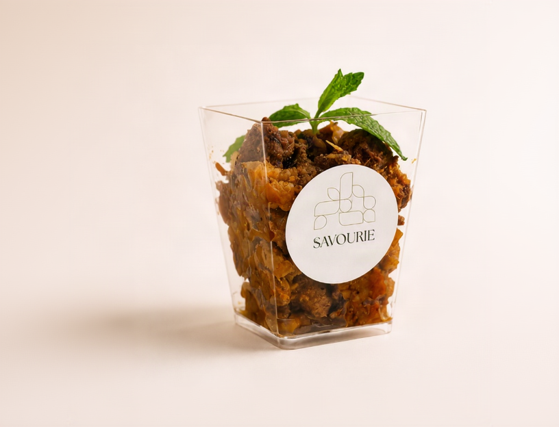 A clear square plastic cup filled with a brown crumbly dessert or snack, topped with a green mint leaf, on a plain warm beige background. The cup has a white round label reading 'SAVOURIE'.