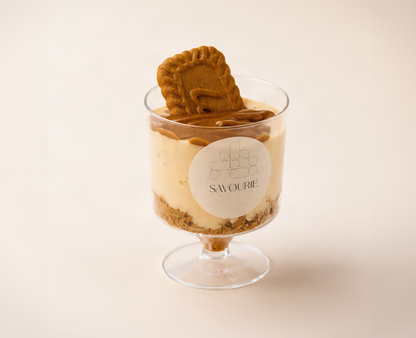 A dessert parfait in a clear glass pedestal cup with a round white label reading 'SAWOURI', layered with cream, chocolate sauce, biscuit, and a cookie on top, set against a warm cream background.