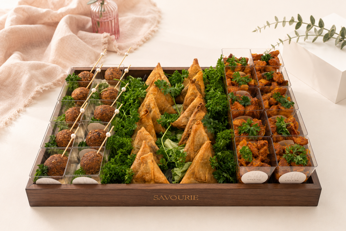 A wooden catering tray labeled 'SAVOURIE' filled with assorted finger foods, including samosas, mini meatballs on skewers, and stuffed vegetables, arranged on a light beige surface with soft decorative props.
