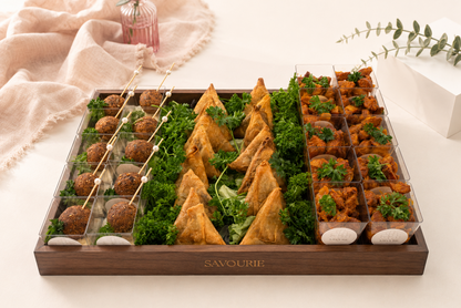 A wooden catering tray labeled 'SAVOURIE' filled with assorted finger foods, including samosas, mini meatballs on skewers, and stuffed vegetables, arranged on a light beige surface with soft decorative props.