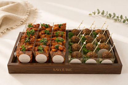 A wooden catering tray labeled 'SAVOURIE' holding two sections of appetizers: clear plastic cups filled with orange-red fried bites topped with parsley, and clear cups filled with brown ball-shaped snacks on toothpicks, set on a light beige surface with a draped fabric and eucalyptus sprig.