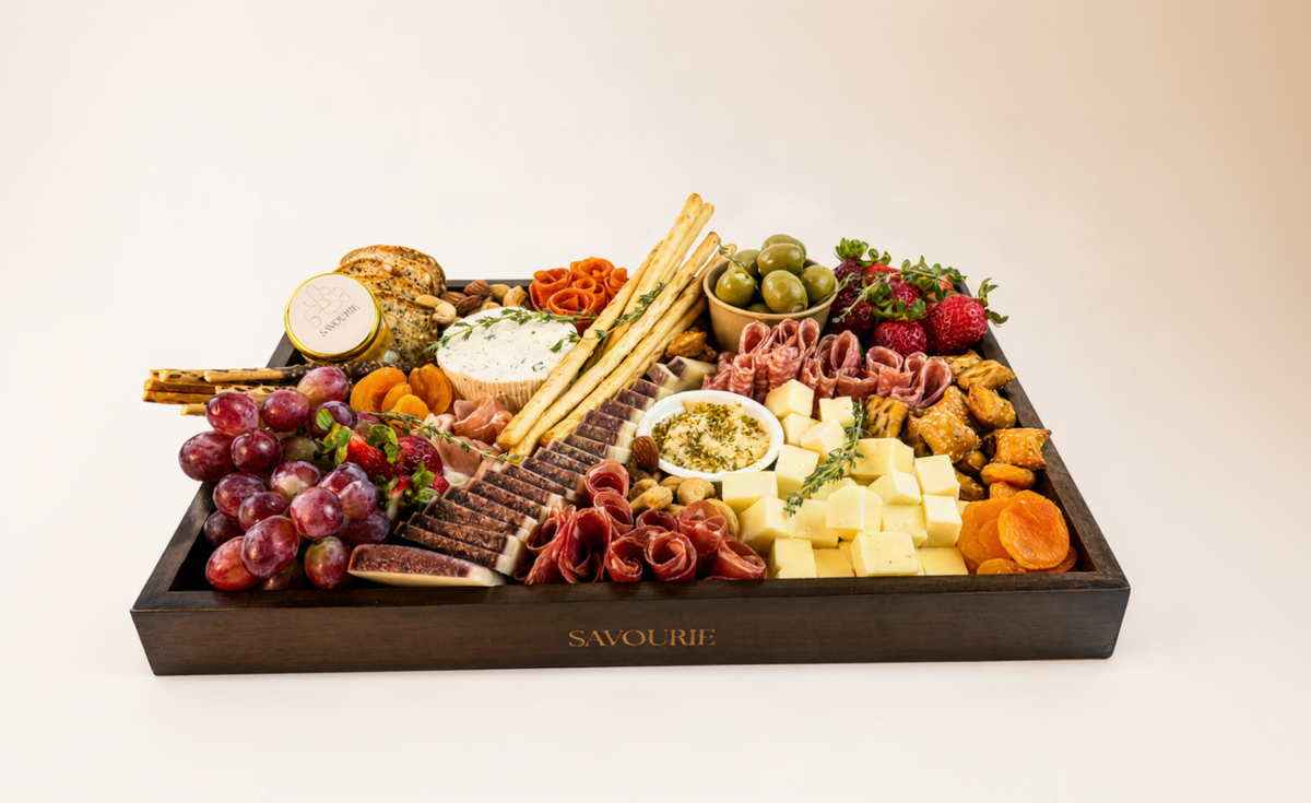 A gourmet charcuterie board in a dark wooden tray branded 'SAVOUR', filled with cheeses, cured meats, crackers, breadsticks, olives, grapes, strawberries, and fruit, photographed on a clean white background.
